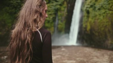 Uzun saçlı bir kadın Endonezya 'daki güçlü bir şelalenin önünde zarif bir şekilde duruyor, yavaş çekim görüntüleriyle..