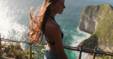 Bikinili bir kadın, denizi gören kayalık bir uçurumda güven içinde duruyor. Uçurum, Endonezya 'nın Nusa Penida Adası' ndaki Kelingking Gizli Nokta Sahili 'nde yer almaktadır..