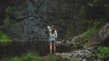 Genç bir kadın gür bir ormanda şelale havuzunun yanında duruyor, uzun saçlarına dokunuyor, Fransız Polinezya Adası 'nın huzurlu manzarasının tadını çıkarıyor.