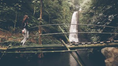 Endonezya, Bali 'deki görkemli bir şelalenin üzerinde asılmış bir köprüden zarif bir şekilde geçen bir kadın. Şelale gibi akan su, yolculuğuna büyüleyici bir zemin hazırlar..