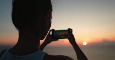 Kadın silueti Tayland 'da dinlenirken telefonuyla gün batımının güzel bir fotoğrafını çekiyor. Kız parlak renkli gökyüzünün tadını çıkarıyor. Açık hava yaşam tarzı seyahati, yaz tatili, ağır çekim. Kapat.
