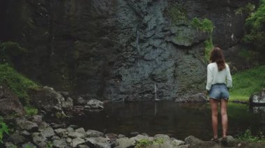 Yalnız bir kadın, Fransız Polinezyası 'nın verimli tropikal manzarasında çağlayan bir şelaleye hayran bir şekilde kayalık bir sahilde duruyor. Su yeşil bitkilerle çevrili sakin bir havuza akar..