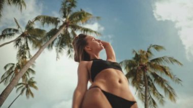 Bikinili bir kadın Tayland 'da, sıcak güneşin altında uzun palmiye ağaçlarının önünde zarif bir şekilde duruyor. Sahne tropikal titreşimler ve rahatlama yayar..