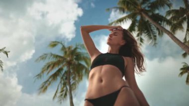 Genç bir kadın tropikal bir plajın tadını çıkarıyor, sıcak güneşi hissediyor, deniz ve gökyüzünün nefes kesici manzarasını çekiyor, rüzgarda sallanan palmiyelerle çevrili.