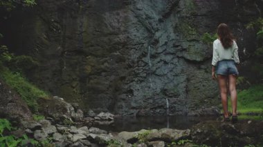 Yeşillik ve sükunetle çevrili gür bir ormanda huzurlu bir şelalenin tadını çıkaran genç bir kadın. Fransız Polinezyasındaki güzellik deneyimi.