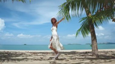 Beyaz elbiseli ve şapkalı bir kadın Tayland 'daki beyaz kumsalda yürüyor. Arka planda palmiye ağacının gölgesinde ve okyanusta yürüyen bir kız. Kadın turist yaz tatili. Yaşam tarzı tatil konsepti