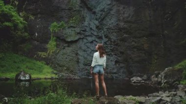 Genç kadın sakin dağ göletinin kenarında duruyor, yemyeşil orman ve şelaleli kayalık dağ duvarı. Kız yaz tatilinde açık hava seyahatinde rahatlar. Dramatik karanlık panorama