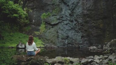 Genç bir kadın, Fransız Polinezyası 'ndaki yemyeşil bir şelalenin önünde, bir kayanın üzerinde bağdaş kurarak meditasyon yapıyor. Tropikal manzara, küçük su havuzuyla sakin ve huzurludur. Arka plan, koyu tonlama