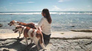 Güneşli bir günde köpeğini okyanusun kenarında okşayan kadın esintiyi hissediyor ve doğa güzelliğiyle çevrili dalgaları dinliyor.
