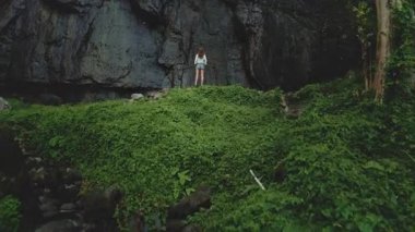 Gökyüzü manzaralı turist kadın yüksek kaya şelalesine karşı yeşil tropikal orman ormanında duruyor. Yaz tatilinde açık hava seyahati. Dramatik koyu tonlama. İHA atışı yükseğe yakınlaştır