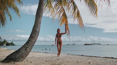 Bikinili genç bir kadın tropikal kumsalda yürüyor, hindistan cevizi palmiyesi dallarıyla oynuyor. Okyanusta lüks yatlar. Egzotik bir adada rahatla, doğa yaz manzarası. Seyahat, turizm, tatil, tatil