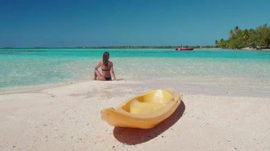 Tropikal ada Bora Bora cennet plaj manzarası. Kadın, beyaz kumlara, sarı kanoya, mavi gökyüzüne, kristal suya oturmuş rahatlıyor. Egzotik adada seksi bir kadın. Yaz tatili seyahati, aktif spor