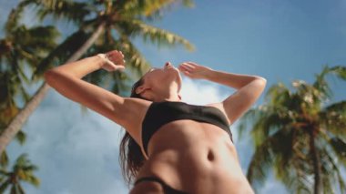 Bikini giyen bir kadın bir sıra uzun palmiye ağacının önünde güvenle duruyor. Açık mavi gökyüzü ve güneşli hava tropikal plaj ortamını güçlendirir..