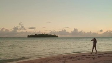 Bir kadın uzak bir adaya doğru uzanan sakin mavi sularıyla Maldivler 'de el değmemiş bir plaj boyunca yürüyor. Gökyüzü yumuşak bir pastel pembe ve turuncu, güzel bir günbatımını gösteriyor..