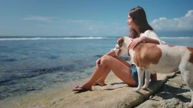 Bir kadın büyük bir kayanın üzerinde küçük bir köpeğin yanında oturuyor. Rahatlamış görünüyor ve önündekilere bakıyor. Köpek onun yanında oturuyor, ona bakıyor..