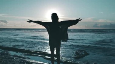 Kadın gün batımında deniz sahilinde el kaldırıyor. Okyanus kıyısındaki kadın silueti, rüzgar saçı ve elbisesi. Mavi gökyüzü, güneş ışığı suya yansıyor. Rahatla, yaz tatilinin tadını çıkar. Arka plan yavaş çekim