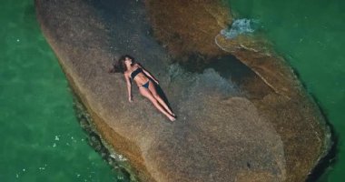 Siyah bikinili genç bir kadın canlı turkuaz okyanus suyuyla çevrili büyük bir kayanın üzerinde uzanır. Hava aracı atışı. Kadın dinleniyor ve güneşleniyor. Tatil seyahati kavramı