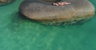 Kadın, berrak turkuaz sularla çevrili pürüzsüz bir kayanın üzerinde güneşlenerek sakin sahil atmosferini geliştiriyor. Güneş tepemizde ışıl ışıl parlıyor, rahatlamak için mükemmel bir gün yaratıyor..