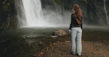 Dişi yürüyüşçü, bereketli bir tropikal yağmur ormanında kayalık bir uçurumdan yuvarlanan güçlü bir şelaleye hayran. Endonezya 'daki macerası sırasında doğanın huzur ve sükunetinin tadını çıkarıyor.