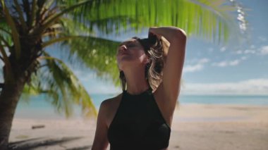 Genç bir kadın tropik bir plajda bir palmiye ağacının altında güneşleniyor ve turkuaz okyanusun tadını çıkarıyor. Cennete benzeyen bir ortamda tasasız ve mutlu bir hayat sürüyor.