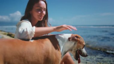 Kadın ve köpeği kumsalda güneşli bir günün tadını çıkarıyorlar, gülümsüyorlar ve sakin atmosferde dinleniyorlar.