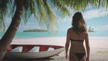 Güzel genç bir kadın tropikal bir sahilde palmiye ağacının altında bir kayıkla yürüyor. Güneşin ve gölün turkuaz suyunun tadını çıkarıyor.