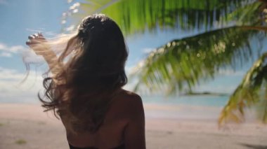 Genç bir kadın, arka planda bir palmiye ağacıyla tropik bir plajda dururken kollarını kaldırıyor. Sıcak havanın ve okyanus esintisinin tadını çıkarırken saçları rüzgarda savruluyor.