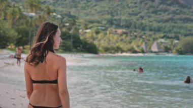 Güneşli bir plaj gününün tadını çıkaran bir kadın, arkasında yeşil tepeler olan okyanusa doğru yürüyor, ılık güneşi ve yumuşak kumu hissediyor, kaygısız bir ruhla güzelliği kucaklıyor.