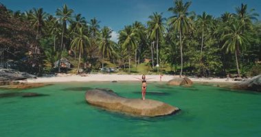 Bir kadın, kollarını açmış, palmiye ağaçları ve turkuaz suyla çevrili güzel bir sahilde bir kayanın üzerinde duruyor. Güneşli bir gün, rahatlamak ve eğlenmek için mükemmel..