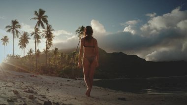 Fransız Polinezyası 'nda beyaz bir kumsalda çıplak ayakla yürüyen bir kadın. Arka planda palmiye ağaçları ve dağ var. Güneş batıyor, sahnede altın bir parıltı yayıyor..