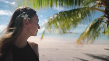 Genç bir kadın tropik bir adada esintinin tadını çıkarıyor, beyaz kumsalda duruyor ve palmiye ağacının altındaki turkuaz okyanus suyuna bakıyor.