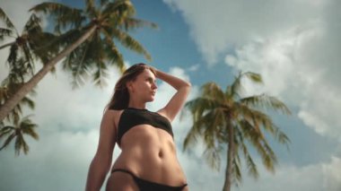 Genç bir kadın tropikal bir plajda duruyor, ılık güneşin ve sallanan palmiye ağaçlarıyla çevrili hafif esintilerin tadını çıkarıyor.