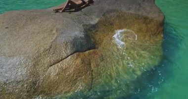 Bir insan, sersemletici turkuaz okyanusun sıcak güneş ışığının tadını çıkararak büyük bir kayanın üzerinde yaslanıyor. Huzur ortamı rahatlamak için mükemmel bir ortam yaratıyor..