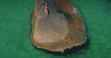 Bir insan, berrak turkuaz sularla çevrili büyük bir kayanın üzerinde uzanarak huzurlu bir anın tadını çıkarır. Bu huzurlu ortam, sahil kenarında geçirilen yaz tatilinin özünü yansıtıyor..