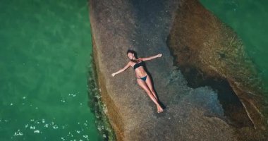 Bir kadın turkuaz suyla çevrili düz bir kaya yüzeyinde yatıyor. Tropikal bir plaj ortamında huzurlu bir anın tadını çıkarırken güneş ışığı canlı renkleri artırıyor..