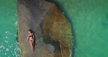 Bir insan, berrak turkuaz su ve sakin bir atmosferle çevrili büyük bir kayanın üzerinde uzanırken güneşin sıcaklığından keyif alır. Parlak yaz günü mükemmel bir rahatlama kaçamağı yaratır..