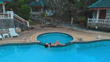 Yuvarlak bir yüzme havuzunun kenarında turkuaz suyla rahatlayan bikinili genç bir kadının hava aracı görüntüsü havuz kenarında bir kedi heykeli görülüyor.