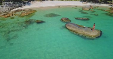 Bir kadın, turkuaz su ve kumlu plajla çevrili bir kayanın üzerinde durmaktan zevk alır. Bereketli yeşillik çarpıcı kıyı manzarasını çerçeveler ve cennet gibi bir ortam yaratır..