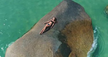 Bir insan, canlı turkuaz suyla çevrili büyük bir kayanın üzerinde güneşlenmekten hoşlanır. Sahne, sakin bir atmosferi sergiliyor. Sıcak bir günde rahatlamak için mükemmel. Doğal güzellikleri vurguluyor..