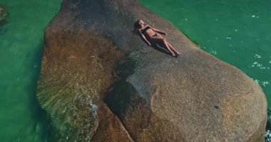 Sıcak güneşin altında yumuşak bir kaya formasyonunda dinlenen bir kadın, etrafındaki turkuaz suların sükunetinin tadını çıkarıyor. Bu huzurlu an, tropikal bir yerde mükemmel bir gün yakalar..