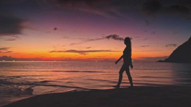 Genç bir kadının silueti kollarını açıyor, özgürlüğün tadını çıkarıyor ve Tayland 'da sakin deniz suyu ve uzaktaki bazı teknelerle tropik plajda güneşli bir gün batımı yaşıyor.