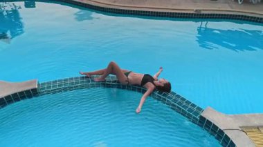 Bikini giyen genç bir kadın günbatımında güzel tropik plajlara bakan sonsuz havuzda dinleniyor, lüks tatil beldelerinde yaz tatilinin tadını çıkarıyor.