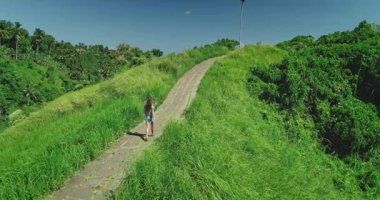 Genç kadın turist, Ubud, Bali 'deki Campuhan Tepesi Yürüyüşü' nde yemyeşil bitki örtüsüyle çevrili asfalt bir yolda yürüyor ve manzaranın tadını çıkarıyor.
