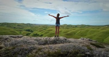 Özgürlüğü kutlayan ve Endonezya 'nın Sumba Adası' ndaki yeşil tepelerin üzerindeki güzel manzaranın tadını çıkaran genç bir kadının arka görüntüsü.