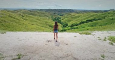 Tepenin tepesinde duran kadın turist, doğa severler ve maceracılar için popüler bir yer olan Sumba, Endonezya 'daki sarp yeşil tepelerin ve yemyeşil bitkilerin çarpıcı manzarasının tadını çıkarıyor.