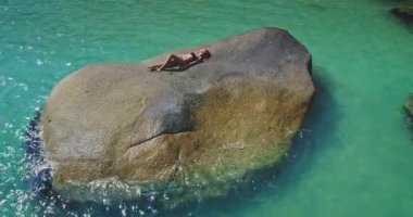 Bir insan el değmemiş turkuaz suyla çevrili büyük bir kayanın üzerinde huzur içinde yatar. Sahne huzurlu bir anı yakalıyor, muhtemelen sıcak ve güneşli bir günde..
