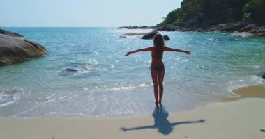 Bir kadın kıyıda kollarını açmış, sakin okyanus esintisini ve güneş ışığını kucaklıyor. Temiz sular ve yumuşak kum, sakin bir atmosfer yaratır..