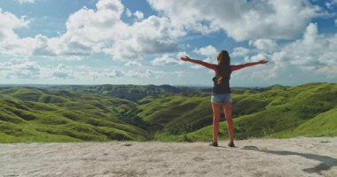 Endonezya 'nın Sumba Adası' nda güneşli bir yaz gününde kolları açık, yuvarlanan yeşil tepelerin ve mavi gökyüzünün büyüleyici manzarasını kucaklayan kadın turist.