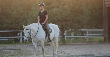 Ata binme eğitimi, genç jokey gün batımında ahırlarda beyaz ata biniyor, binicilik sporu, aktif yaşam tarzı, atları sevmek, doğada boş vakitlerin tadını çıkarmak.