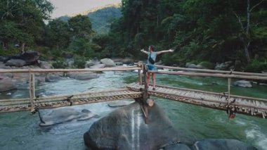 Genç kadın turist, bambu köprüsünde kollarını açmış tropik bir ormandan akan vahşi bir nehrin üzerinde doğanın özgürlüğünün ve güzelliğinin tadını çıkarıyor.